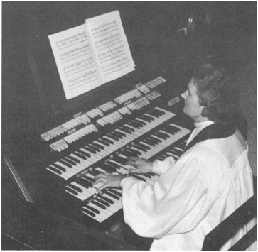 A person in robes plays a large pipe organ with multiple keyboards and visible sheet music, focusing intently on the instrument.