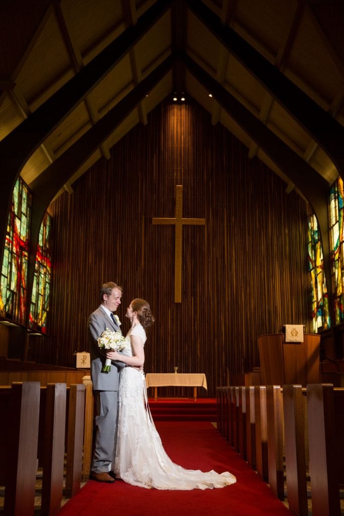 Two people stand in a warmly lit church, under a large wooden cross, beside colorful stained glass, on a red carpeted aisle.