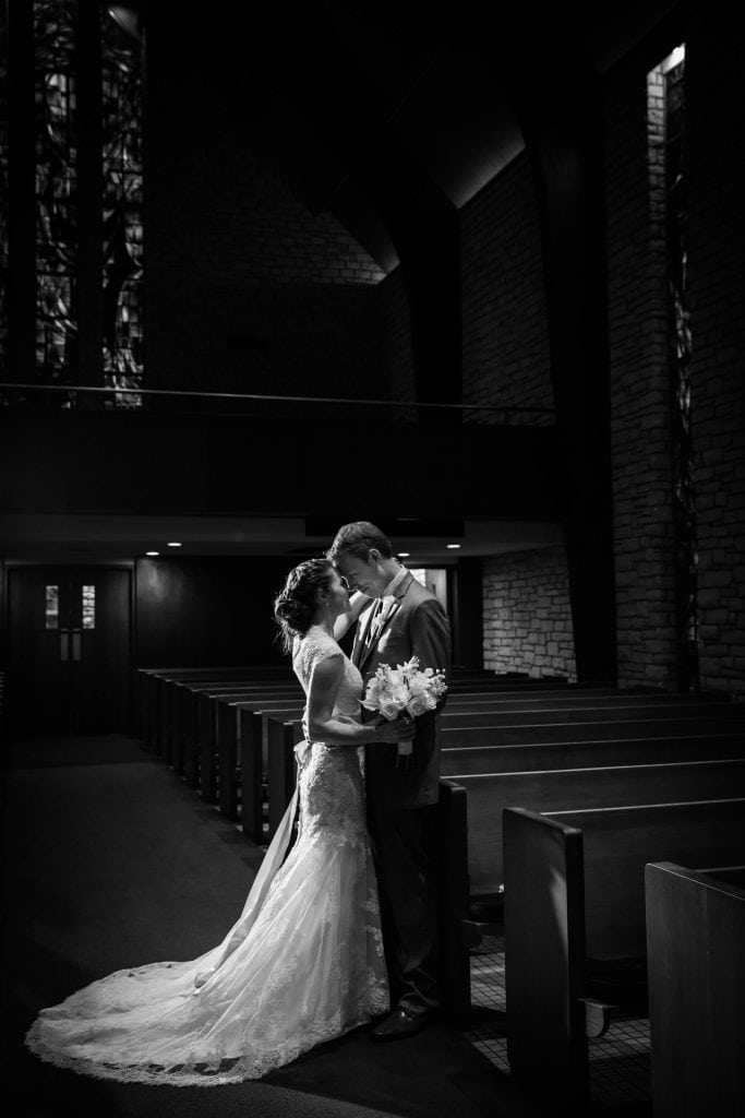 A couple stands closely in a dimly-lit church interior, surrounded by pews, the person in a wedding dress holding a bouquet.