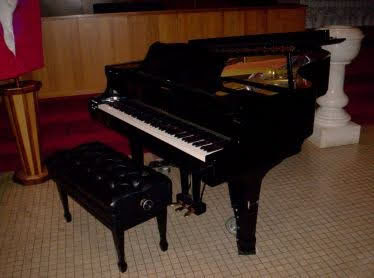 A grand piano with an open lid is situated indoors, accompanied by a cushioned bench on a tiled floor in a formal setting.