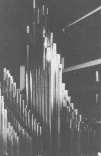 The image shows a close-up of large, vertical organ pipes in a dimly lit space, casting dramatic shadows on surrounding surfaces.