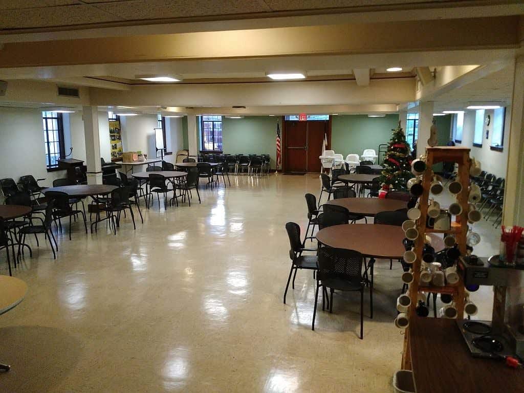 SmootHall A spacious room featuring round tables, chairs, and a Christmas tree. Walls display various items. No people are present.